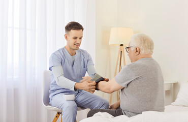 Obraz premium Caring male nurse helping senior patient sitting on bed to put on cuff of blood pressure monitor before routine medical checkup. Hypertension, hypotension, cardiovascular diseases control concept.