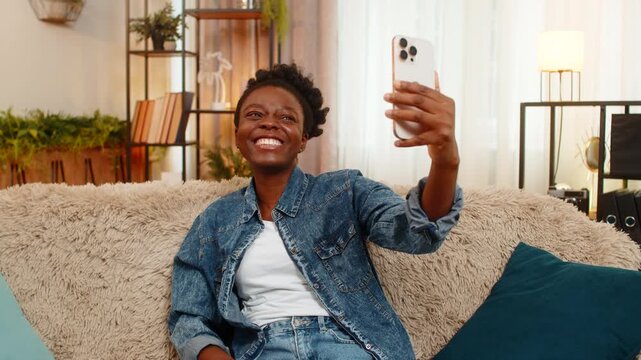 African American young woman smiles joyfully while taking selfies on smartphone, trying different angles, expressions. Black girl enjoys the playful process of capturing trendy shots for social media.