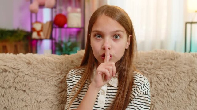 Young child girl sits alone on the sofa and shows a quiet gesture by putting one finger to lips. School kid demonstrates silence signal, looking thoughtful and calm while staying indoors at home.