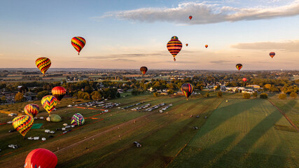 Hot air balloons of various colors soar in the evening sky during a festival in the countryside. The landscape is vibrant with green fields and distant hills under a warm sunset.
