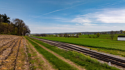Bright blue sky highlights the railway tracks stretching through lush green fields. Trees line the...