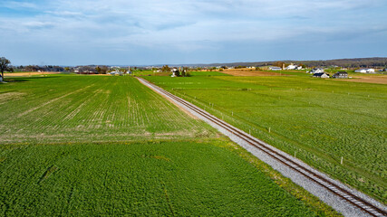 A railway track runs through lush green fields under a clear blue sky. Nearby, small farms and houses dot the landscape, showcasing rural life in a peaceful setting.