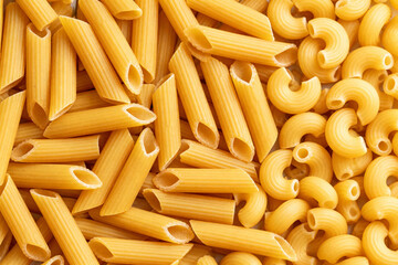 Close up shot of uncooked penne pasta and elbow macaroni pasta scattered on a flat surface