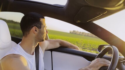 Profile of young handsome man driving a modern electric car with sunlight at background. Young male driver riding on electrical vehicle at countryside. Concept of journey and trip. Close up