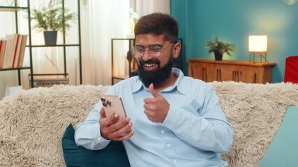 Indian man at home scrolls smartphone news feed smiles as liked post appears enjoys moment happy instant. Arabian guy on sofa taps like button shows emotion approval connection social media happiness. - Powered by Adobe