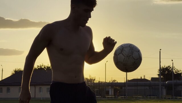 Young man juggling soccer ball on stadium at sunset. Professional footballer kicking ball at green field. Sportsman practicing tricks at meadow with sunlight at background. Freestyle football
