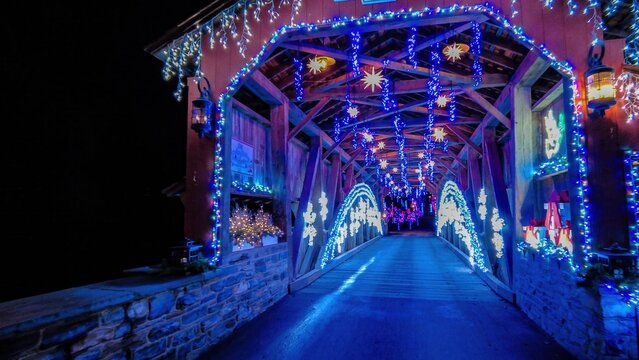 Colorful lights hang from the wooden beams of a covered bridge. The scene is filled with winter decorations, creating a joyful and magical atmosphere during the evening hours of a holiday festival.