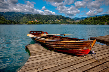 Obraz premium Traditional wooden pletna boats docked along the calm waters of Lake Bled, Slovenia, surrounded by lush greenery and the Julian Alps in the distance on a serene summer morning. 