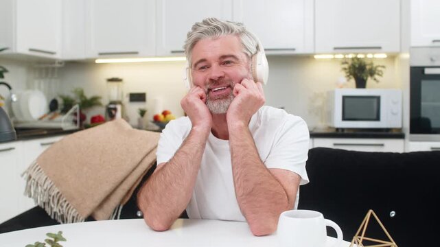 Middle-aged man at home listens to music in wireless headphones leans on hands smiles and dreams. Mature guy at table on sofa thinks about responsible choice future plan feels calm and confident sure.