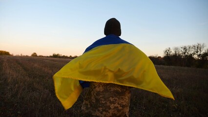 Young male military in uniform jogging with blue-yellow banner on shoulders at meadow. Soldier of...