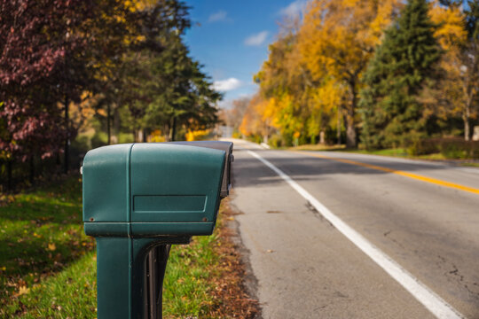 Close-up of a green roadside mailbox on a quiet suburban street during autumn, with colorful trees and soft sunlight in the background. - Powered by Adobe