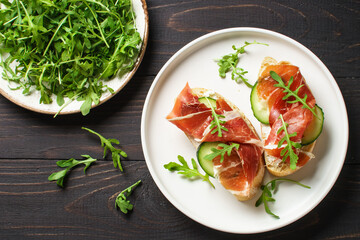 Open sandwiches with cream cheese, prosciutto, cucumber and arugula on white plate.