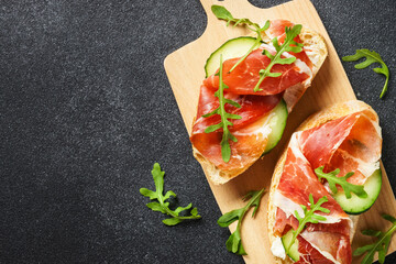 Open sandwiches with cream cheese, prosciutto, cucumber and arugula at cutting board.