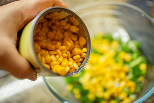 Close-up of a hand pouring vibrant yellow canned corn kernels from a can into a glass bowl of fresh green salad