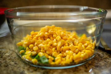 Vibrant yellow corn kernels piled on top of chopped green onions in a clear glass bowl on a brown granite kitchen countertop