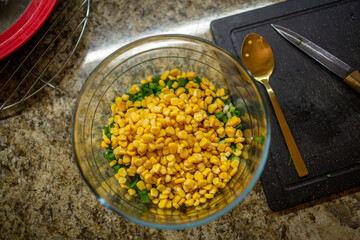 Vibrant yellow corn kernels piled on top of chopped green onions in a clear glass bowl on a brown granite kitchen countertop