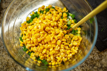 Vibrant yellow corn kernels piled on top of chopped green onions in a clear glass bowl on a brown granite kitchen countertop