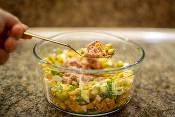 Close-up of a golden spoon lifting a mixture of tuna, corn, and eggs from a glass bowl while preparing a healthy homemade salad