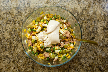Top-down view of creamy mayonnaise being added to a tuna salad with corn, eggs, and pickles in a glass bowl on a granite countertop