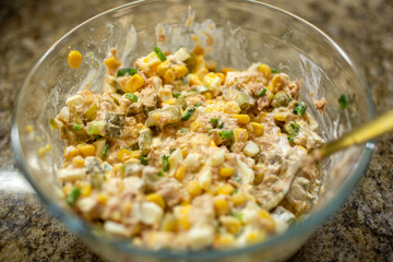 Close-up of a mixed, creamy homemade salad with tuna, corn, eggs, and chopped pickles in a glass bowl on a granite countertop