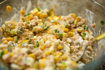 Close-up of a mixed, creamy homemade salad with tuna, corn, eggs, and chopped pickles in a glass bowl on a granite countertop