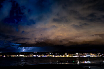 Nighttime shot of a thunderstorm over the coast, with a lightning bolt illuminating the sky above the city. Dark clouds blend with warm and cool tones, while the lights of the port and the city reflec