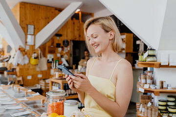 Smiling woman using smartphone buying products at store. Сonscious shopping and retail of organic food and eco goods at local small shop.