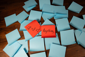Handwritten text reading we can do hard things on red sticky notes in a group of light blue sticky notes positioned diagonally on a dark wood desk.