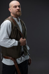 A charismatic bald man in the image of a wealthy medieval inhabitant, in a white blouse and vest, holding a battle axe, tied with a leather belt, photo on gray in the studio