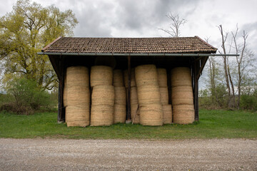 A rustic hut filled with round hay bales stands in a forest meadow. Peaceful rural scene with greenery, natural light, and countryside agricultural elements.