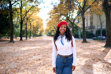 Smiling African millennial woman standing in an autumn park, wearing a beret, scarf and jeans. Natural-light portrait with tree-lined path and golden leaves, warm and relaxed mood.