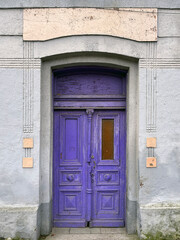 Old wooden double door painted in vibrant purple, set in a gray wall with decorative details and a blank stone sign above, showing weathered texture and vintage architectural style.