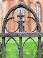 Close-up of an ornate wrought iron fence with a fleur-de-lis style centerpiece and gothic arch patterns, showing rust and weathering, with a blurred historic red brick building in the background.