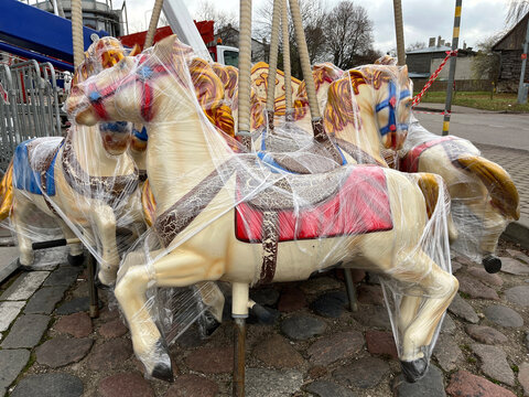 Carousel horses wrapped in protective plastic, standing on cobblestone pavement in an outdoor setting, with colorful saddles and harnesses visible beneath the wrapping.