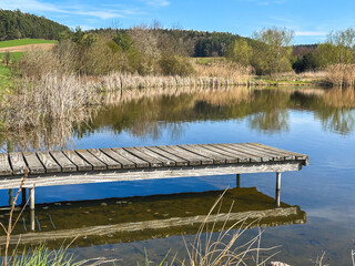 A small wooden dock stretches into a peaceful lake, used for small boats, fishing, or quiet relaxation. Serene water, natural surroundings and soft light create a tranquil outdoor scene.