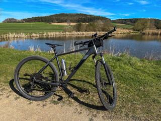 A black bicycle is parked on a cycle path overlooking a calm lake and green forest. Peaceful outdoor scenery ideal for travel, recreation, cycling, and nature themes.