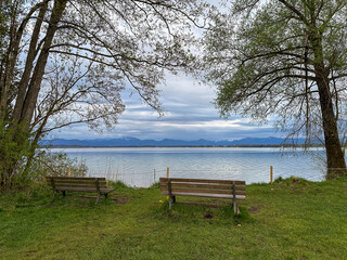 Wooden benches facing a calm lake, offering a peaceful place to rest and enjoy the scenic water view. Soft light, gentle reflections and natural surroundings create a relaxing outdoor atmosphere.
