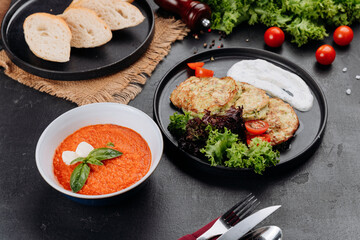 Tomato soup and zucchini fritters with yogurt sauce, salad and bread on dark table