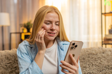 Young woman holding cheek at home, showing tension and urgent need for dental care. Girl scrolling...