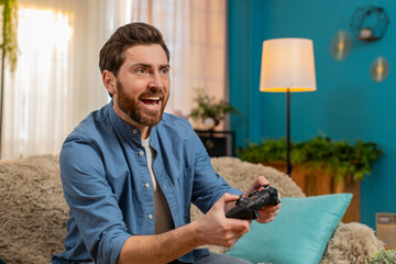 Young man sitting at home on sofa, gripping gamepad joystick, intensely playing video game with focus and excitement. Caucasian guy reacting emotionally to gameplay, immersed in virtual world, indoors