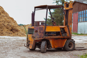 Rusty industrial forklift parked on a gravel surface near a haystack, showcasing weathered textures and machinery in an agricultural setting with rustic buildings