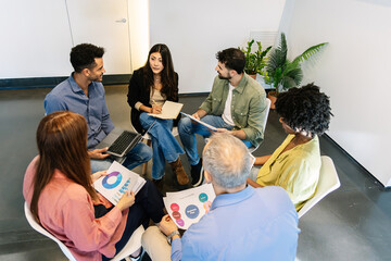 Business group sitting in circle discussing planning ideas. Coworkers reviewing charts in open workspace. Collaboration and planning concept.