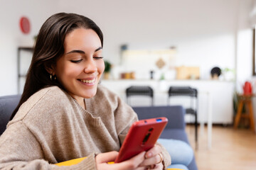 Happy young woman using mobile phone sitting on sofa at home, enjoying cozy moment chatting with friends or watching social media content online. Technology lifestyle concept