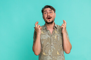 Middle-aged man crossing fingers with tense expression, silently hoping for positive result or good outcome. Young adult guy shows nervous anticipation and emotional hope isolated on blue background.