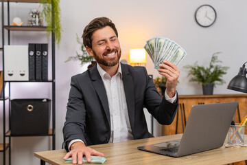Middle-aged businessman at home office counts dollar bills compares numbers with profit chart on laptop surprised. Freelancer guy at table stacks cash pile smiles at gain and plans next investment