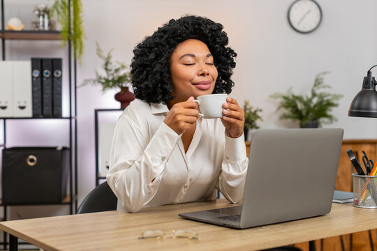 African American businesswoman at home office slowly sips coffee gazing out window letting ideas flow. Freelancer girl at table pauses work enjoying calm drink moment relaxing mind before next task.