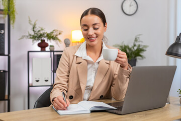 Young businesswoman at home office drinks coffee slowly reads plan in notebook and sets priority list. Freelancer girl at table writes first task opens laptop and begins focused work with calm rhythm