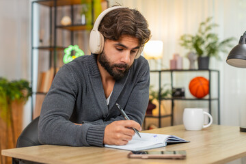 Indian man at home listens to audiobook with headphones takes notes in notebook stays focused....