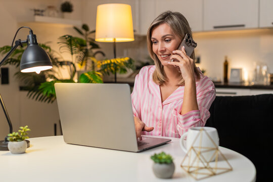 Adult woman at night home works on laptop talks on smartphone manages tasks stays focused. Caucasian girl on sofa at evening at table on sofa speaks on call makes notes replies calm tone focused well.