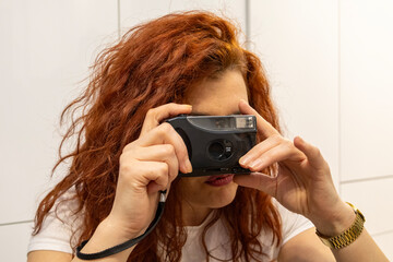 Curly red-haired woman holds a compact retro-style camera to her eye, concentrating on composition; close indoor portrait highlights intent expression and photographic focus in a cozy space.